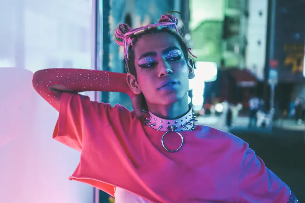 A person wearing a bright pink top and a spiked choker necklace with a large ring, standing in an urban nighttime setting with colourful neon lighting. The background features blurred city lights and street elements, creating a vibrant and edgy atmosphere