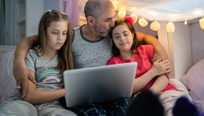 A man sitting between two children on a bed, all looking at a laptop screen together. The setting includes soft lighting with string lights and a cozy blanket fort, suggesting a relaxed, intimate family moment.