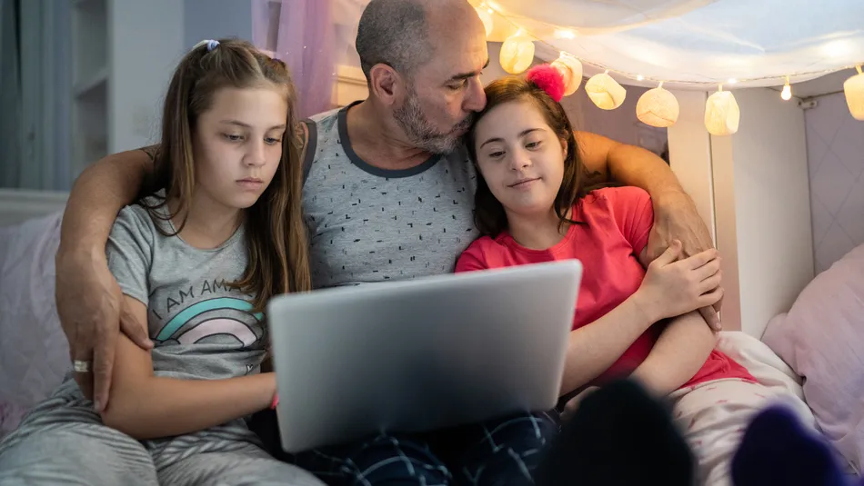 A man sitting between two children on a bed, all looking at a laptop screen together. The setting includes soft lighting with string lights and a cozy blanket fort, suggesting a relaxed, intimate family moment.