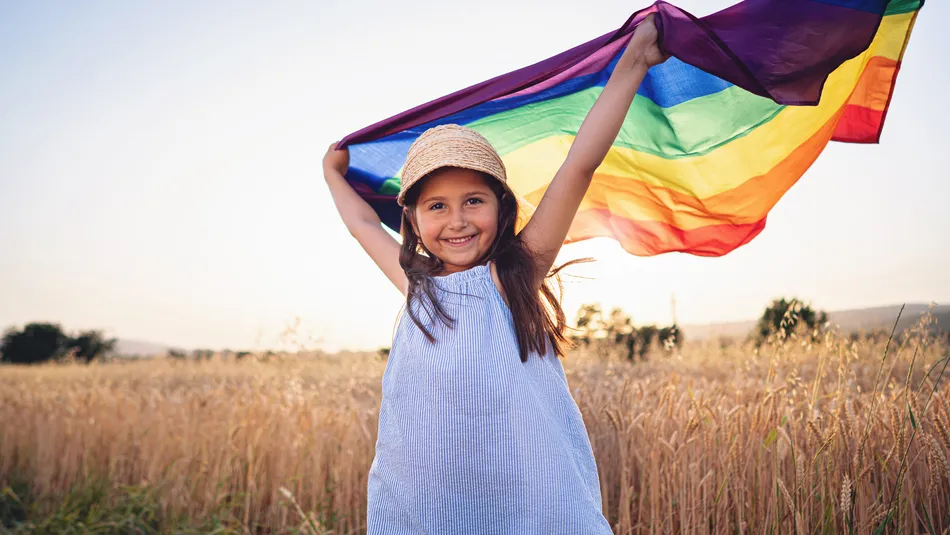 A young girl standing in a wheat field at sunset, holding a rainbow pride flag above her head. The scene highlights themes of freedom and celebration with warm natural lighting and vibrant colours of the flag contrasting against the golden field.