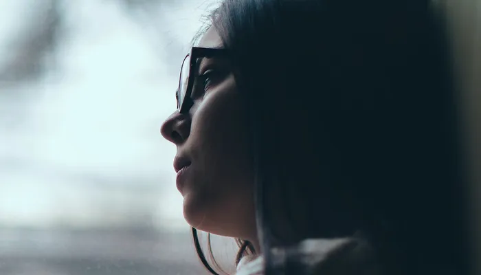 Teenager sitting by a window with arms crossed, looking outside at a blurred urban scene with trees and buildings. The scene conveys a contemplative or reflective mood with soft natural lighting and muted colours.