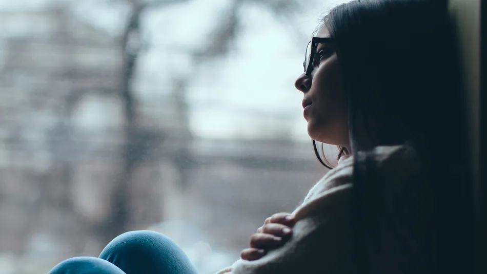 Teenager sitting by a window with arms crossed, looking outside at a blurred urban scene with trees and buildings. The scene conveys a contemplative or reflective mood with soft natural lighting and muted colours.