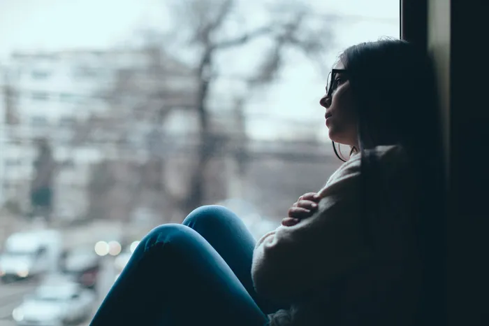 Teenager sitting by a window with arms crossed, looking outside at a blurred urban scene with trees and buildings. The scene conveys a contemplative or reflective mood with soft natural lighting and muted colours.