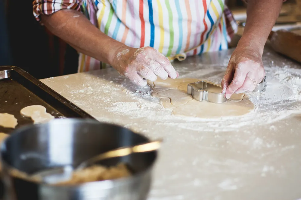 A person using a metal cookie cutter to shape dough on a floured surface, wearing a colourful striped apron. A baking sheet with cut dough shapes and a mixing bowl with a spoon are visible, highlighting a baking preparation process.