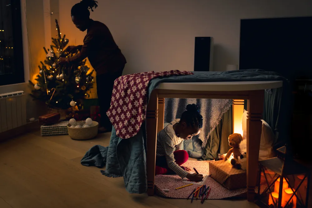 A cozy indoor scene with a child drawing in a den underneath a table which has blankets draped over it and illluminated by warm lights and a teddy bear, while an adult decorates a small Christmas tree nearby.
