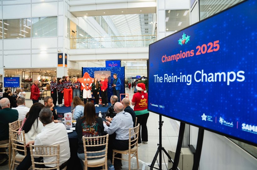 A room of people sitting at round tables playing a quiz with Cosmo, the Action for Children mascot, in the background; a TV screen reads 'Champions 2025: The rein-ing champs'