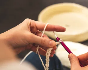 Close-up of hands crocheting with white wool and a purple crochet hook. Background includes a blurred round woven basket, highlighting crafting activity and textile creation.