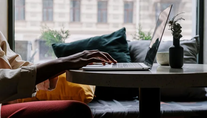 A person typing on a laptop in a cozy indoor setting with a large window showing a blurred building exterior. The scene includes a small vase with a plant on the table and cushions on a bench seat in the background, emphasizing a relaxed work environment