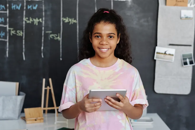 A smiling young girl holding a tablet in a classroom setting with a blackboard behind her displaying a weekly schedule. The scene highlights a modern learning environment combining traditional teaching tools and digital technology.