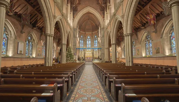 Interior of a large church with rows of wooden pews leading to an ornate altar, stained-glass windows along the walls, and a decorated Christmas tree near the front.