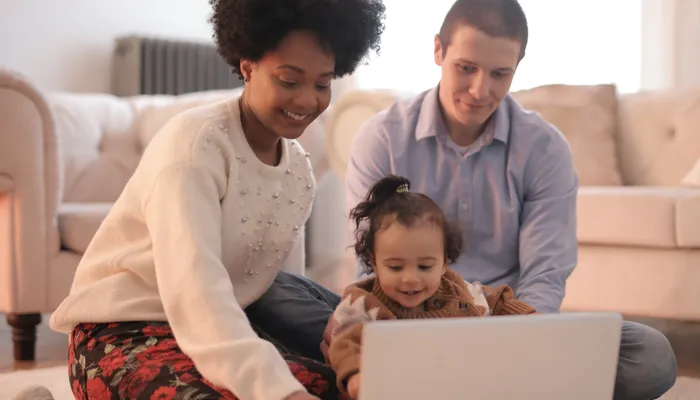 A family sitting on the floor in a living room, engaging with a laptop placed in front of them. The setting includes a beige sofa and neutral-toned decor, highlighting a casual and cozy environment for shared screen time or learning.