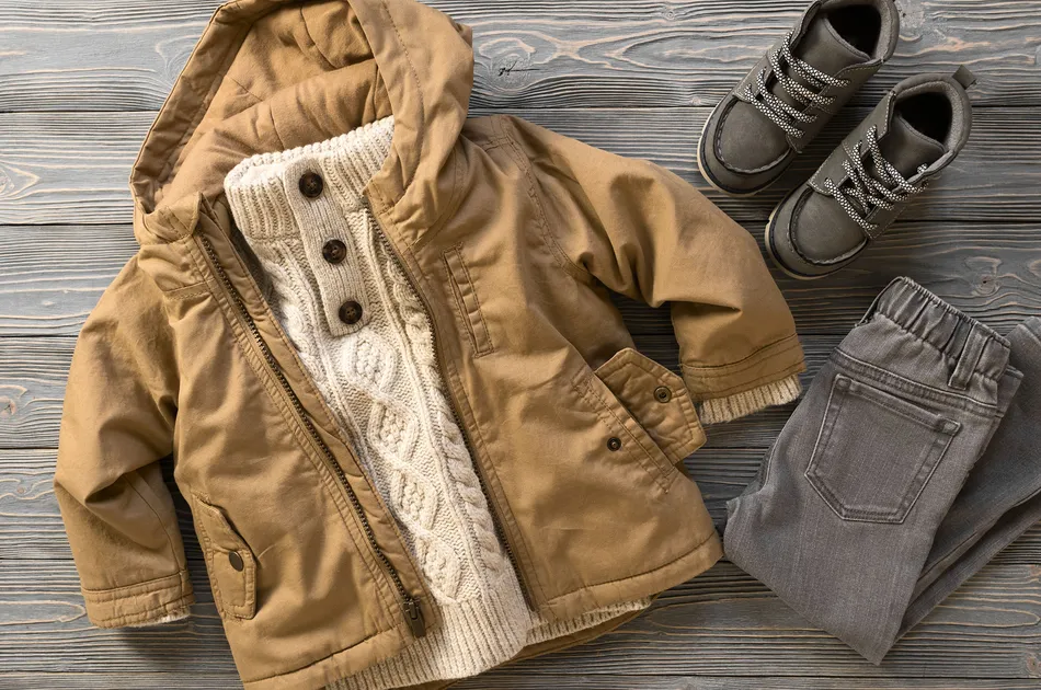 Child's winter outfit laid out on a wooden surface, including a tan hooded jacket, cream cable-knit sweater with buttons, gray trousers, and gray lace-up boots. The clothing items are arranged neatly and show a coordinated cold-weather ensemble