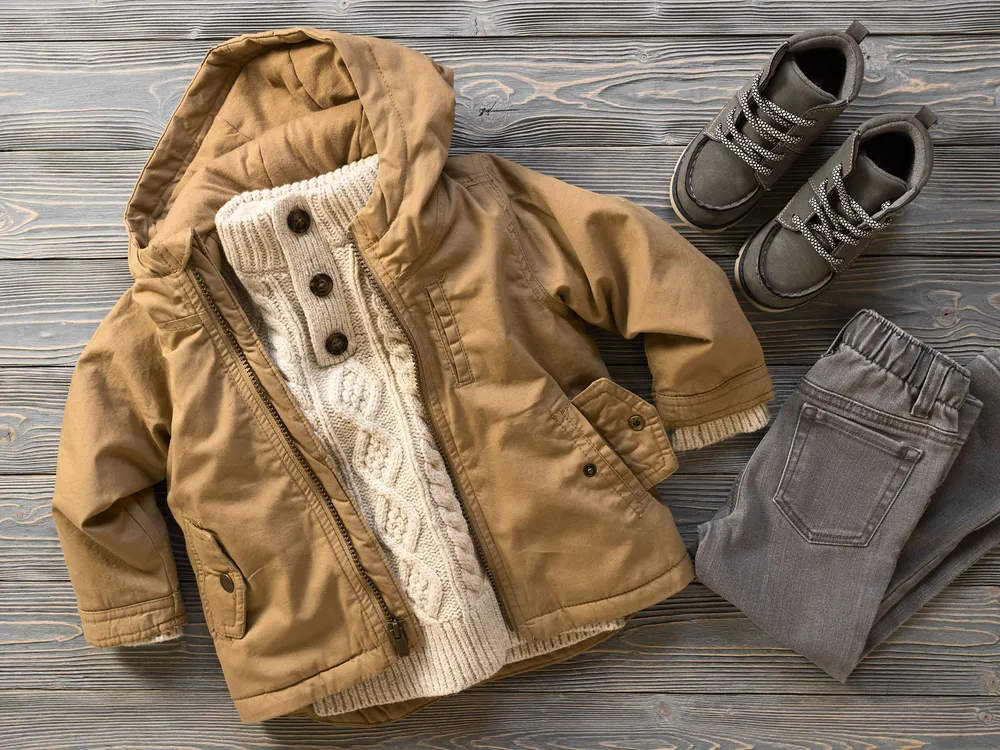 Child's winter outfit laid out on a wooden surface, including a tan hooded jacket, cream cable-knit sweater with buttons, gray trousers, and gray lace-up boots. The clothing items are arranged neatly and show a coordinated cold-weather ensemble