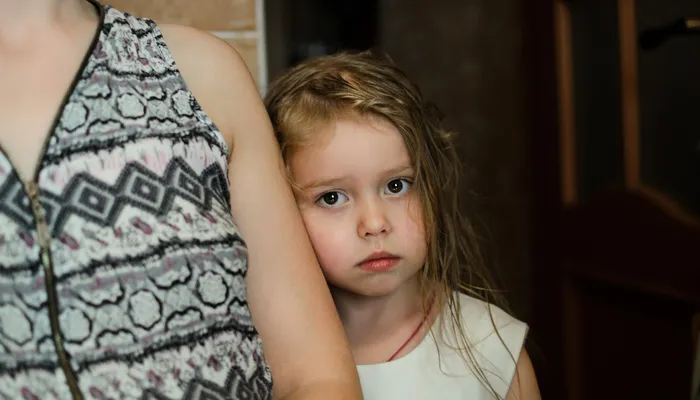 An adult and a sad looking child standing close together. The adult's head is not visible. The setting includes a wooden door and a brick wall in the background.