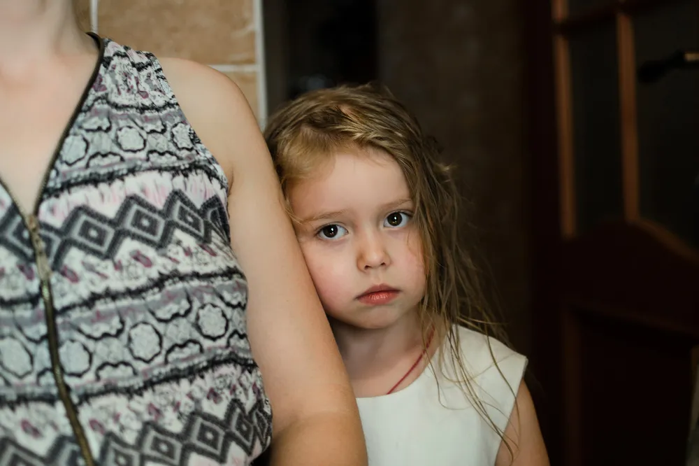 An adult and a sad looking child standing close together. The adult's head is not visible. The setting includes a wooden door and a brick wall in the background.