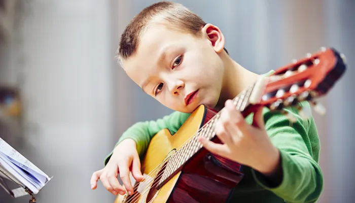 A child playing an acoustic guitar indoors, focusing on hand positioning on strings and fretboard. Music stand with sheet music is visible on the left, indicating a practice or learning session.