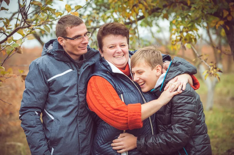 Three people outdoors in a natural setting with autumn foliage, wearing warm jackets in blue, orange, and black colours. The group is closely embracing, suggesting a moment of affection or support among family or friends.