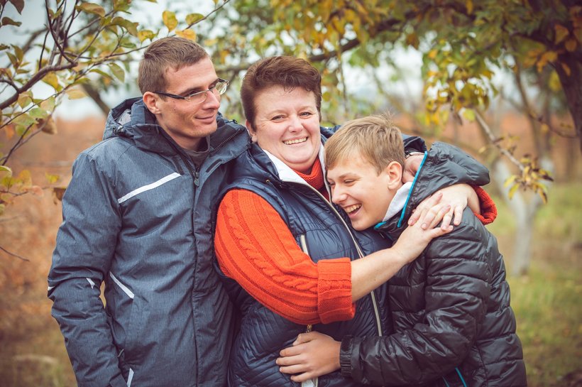 Three people outdoors in a natural setting with autumn foliage, wearing warm jackets in blue, orange, and black colours. The group is closely embracing, suggesting a moment of affection or support among family or friends.