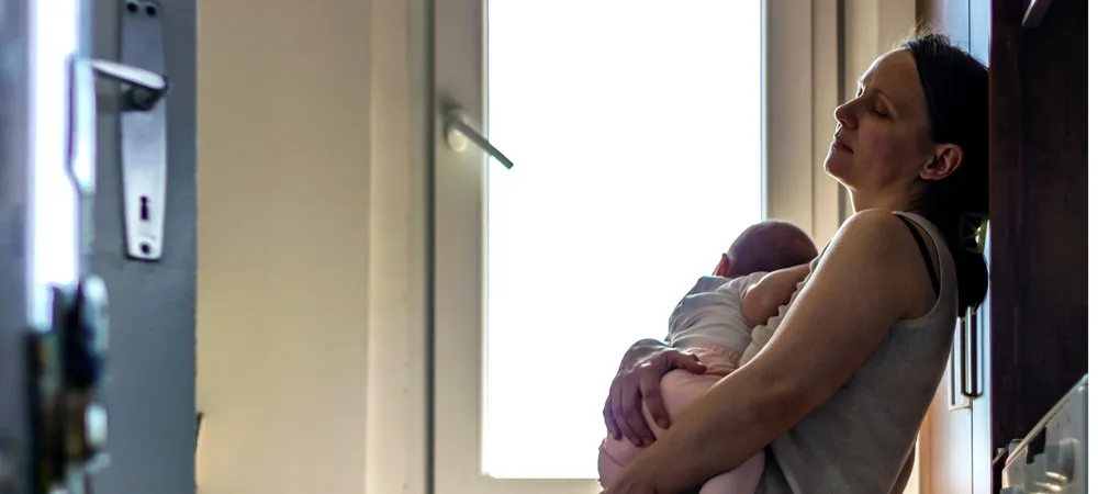 A tired looking woman leaning against a wall near a window holding a baby, conveying a moment of quiet bonding or care. Soft natural light highlights the gentle embrace, with a door and wall framing the scene, emphasizing intimacy and calm.