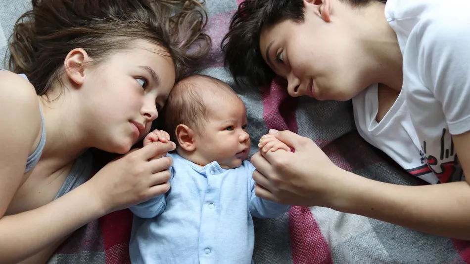 Two children lying on either side of a baby on a checkered blanket, all holding the babies' hands. The scene conveys a sense of family bonding and affection with soft natural lighting and casual clothing.