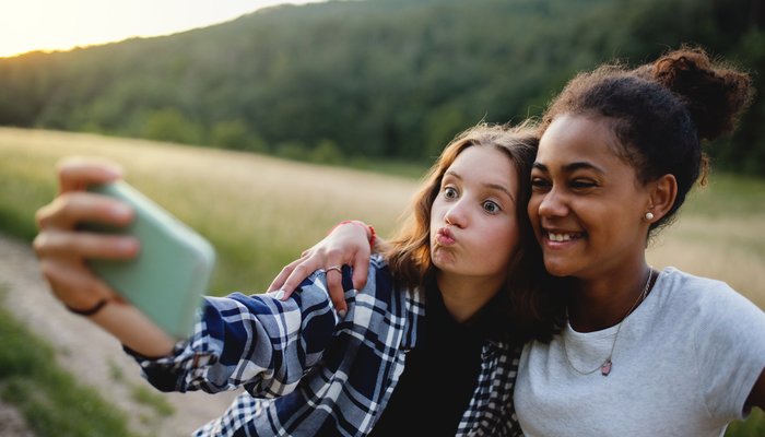 Two young people outdoors taking a selfie with a smartphone during sunset, featuring a blurred background of grassy fields and forested hills. One person wears a checked shirt while the other wears a grey t-shirt, both smiling and posing closely together.