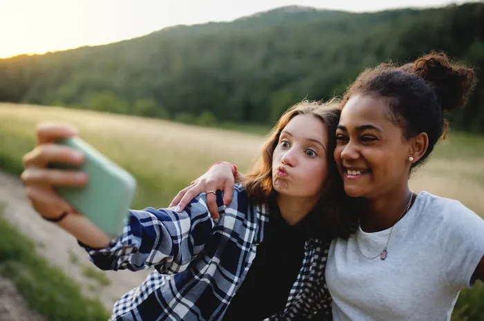 Two young people outdoors taking a selfie with a smartphone during sunset, featuring a blurred background of grassy fields and forested hills. One person wears a checked shirt while the other wears a grey t-shirt, both smiling and posing closely together.