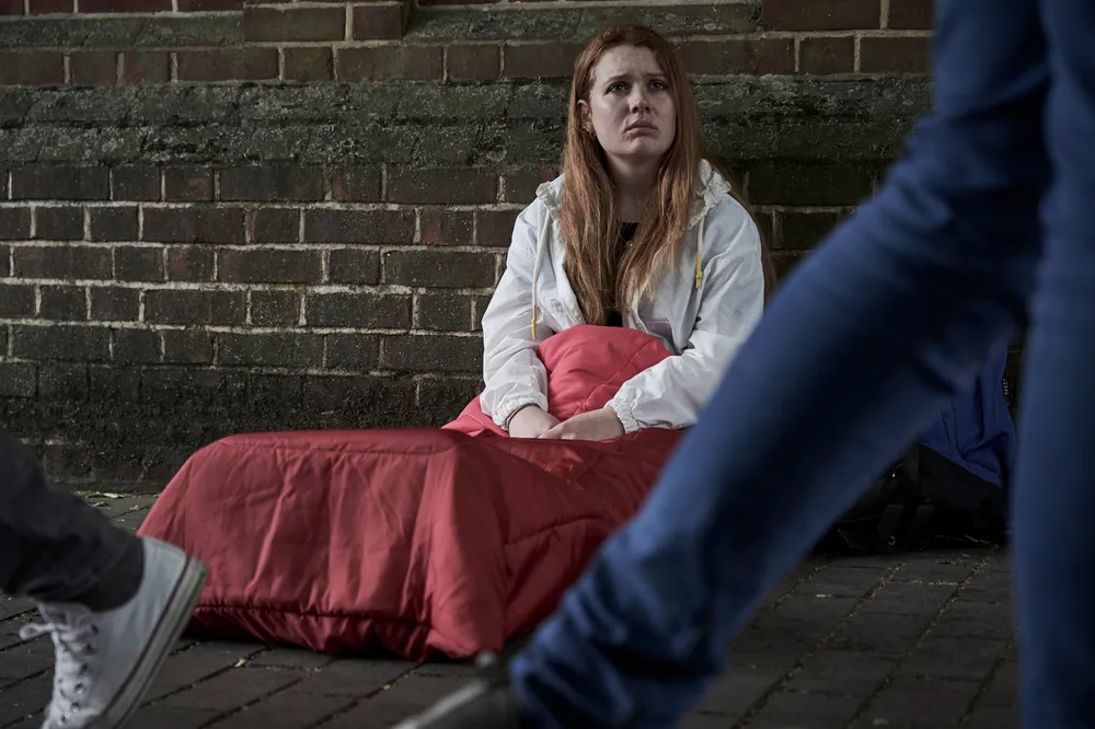 youg homeless girl sitting in sleeping back on street floor as people walk past.jpg