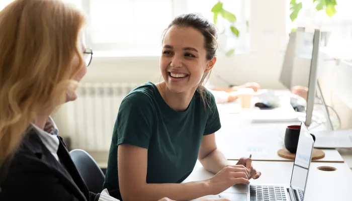 Two people engaged in a conversation at a desk with a laptop and desktop computer. The setting appears to be an office or workspace with natural light and plants in the background.